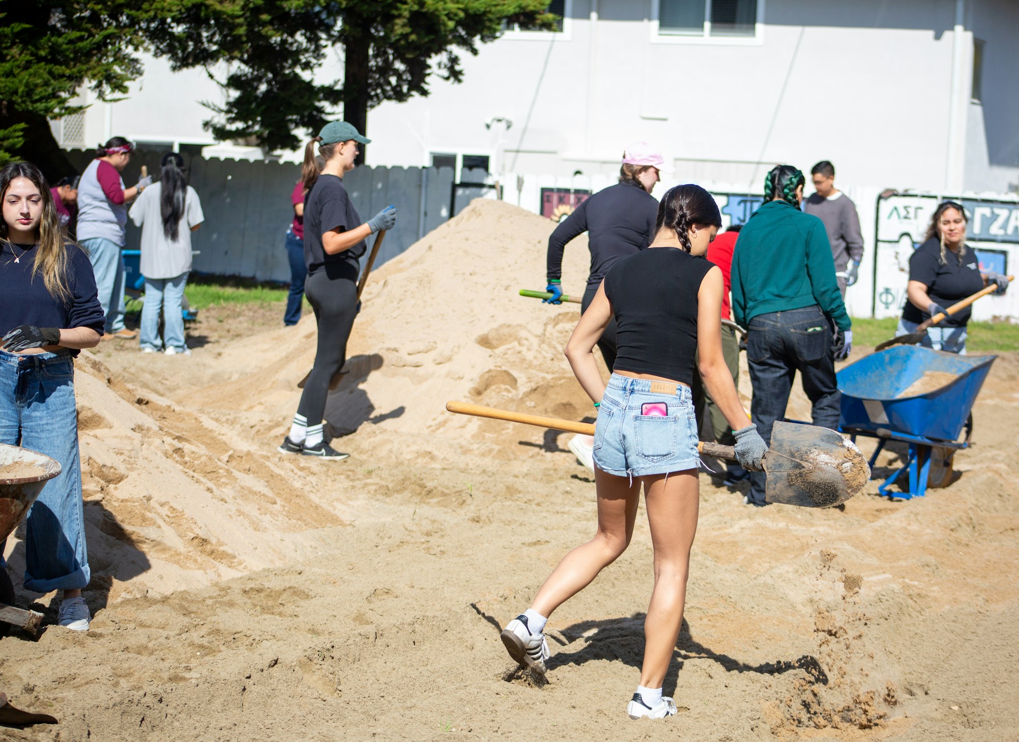 People working together, shoveling dirt or sand into a wheelbarrow at an outdoor site.