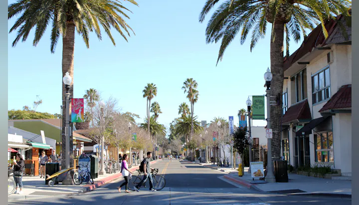 A sunny street scene with palm trees, shops, and people walking with bikes, creating a relaxed atmosphere.