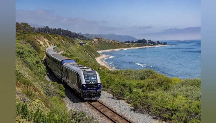 A train runs along a scenic coastal railway, with lush greenery on one side and the ocean on the other under a clear sky.