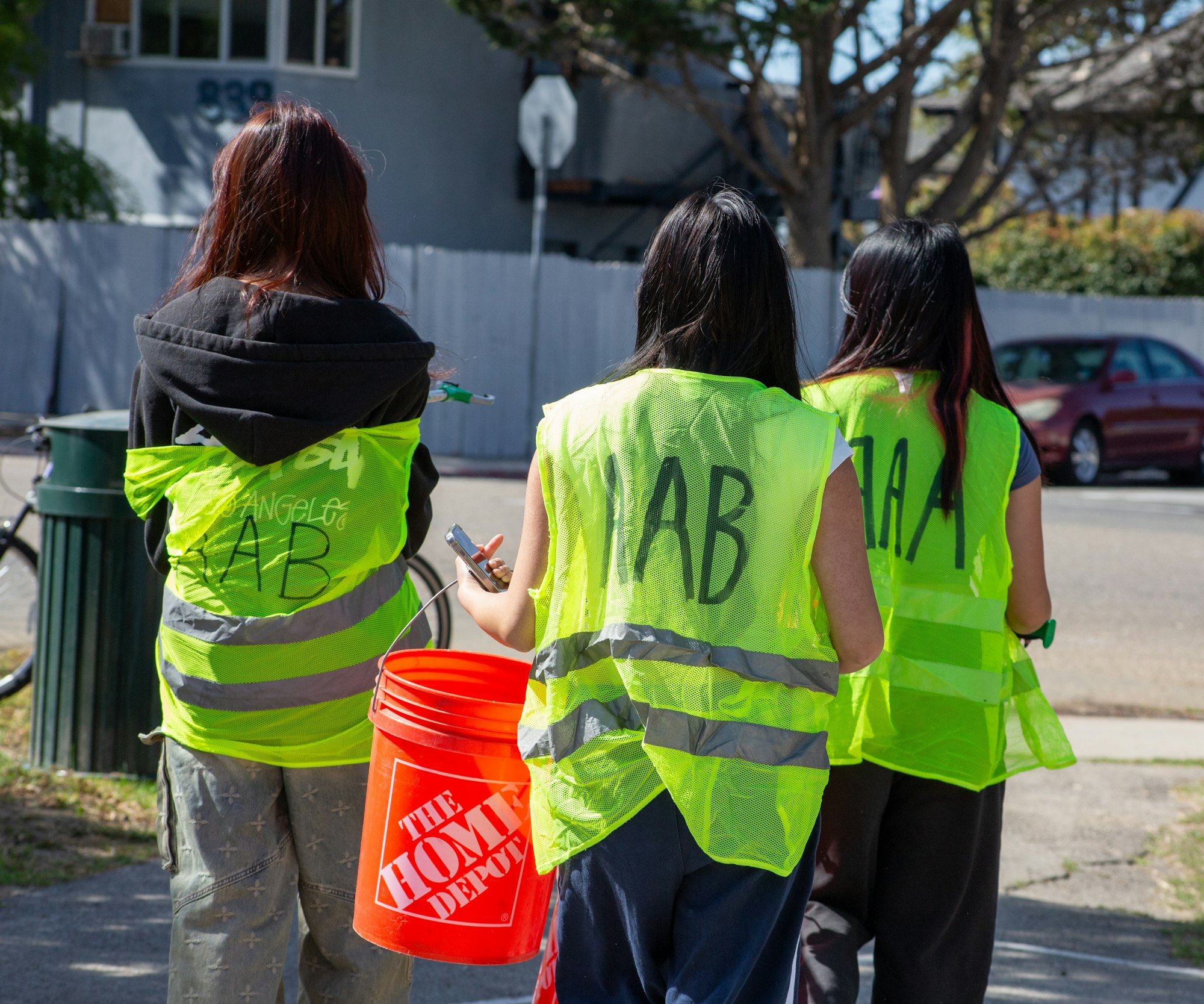 Three people in yellow vests holding an orange bucket walking down a street.