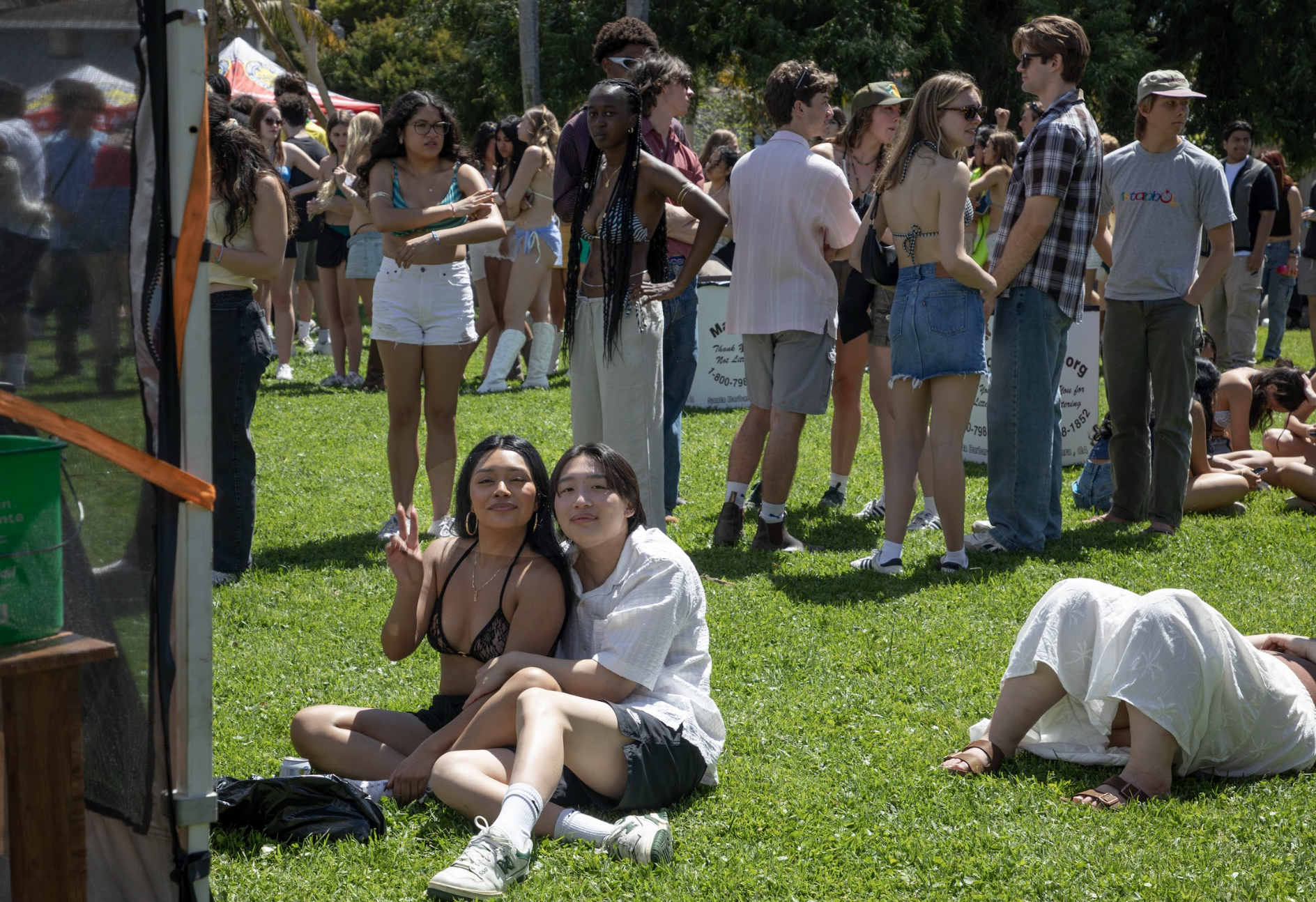 A sunny outdoor event with a crowd, featuring two women posing in front while others socialize and relax on the grass.