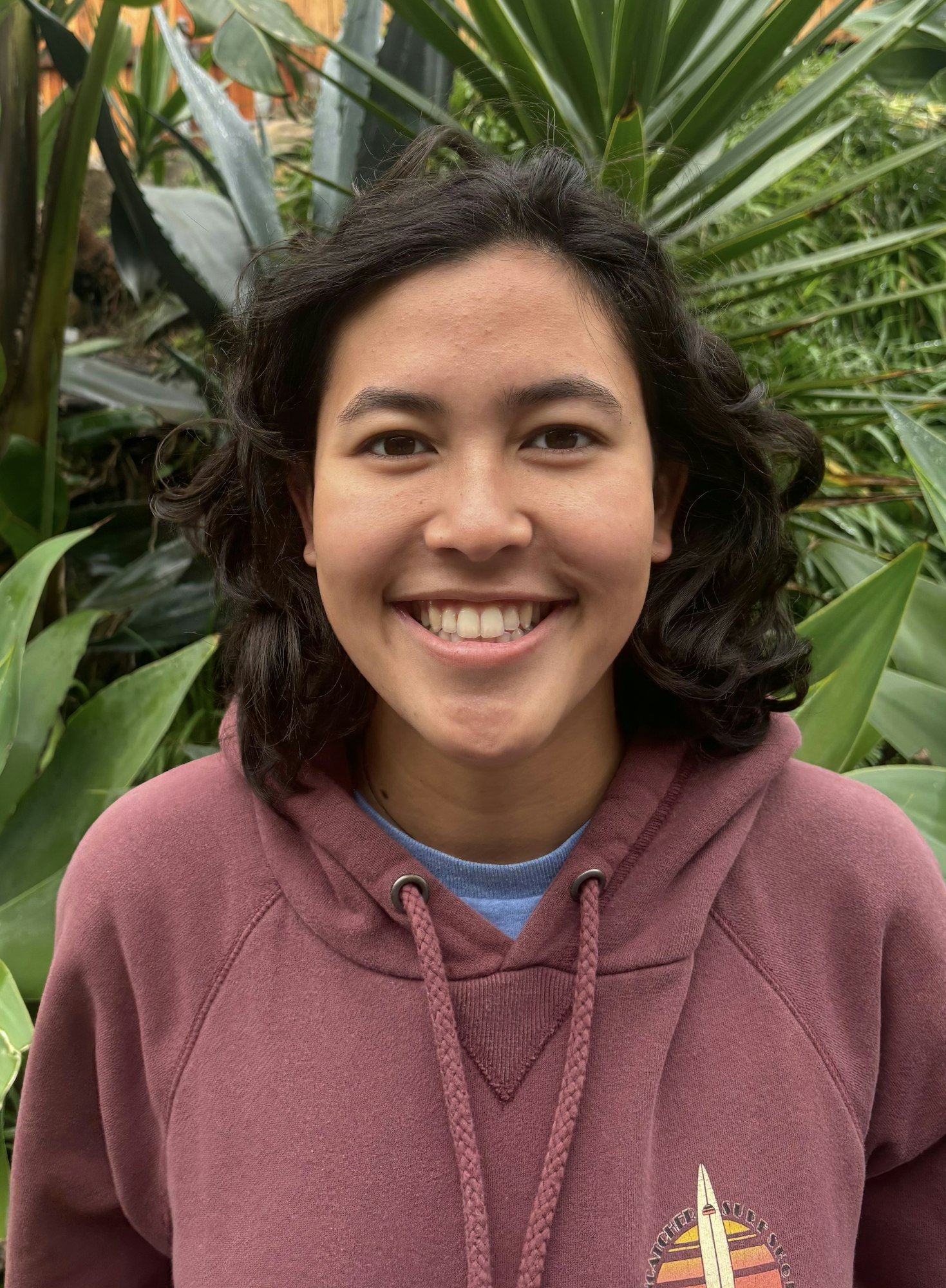 A smiling person in a hoodie with plants in the background.