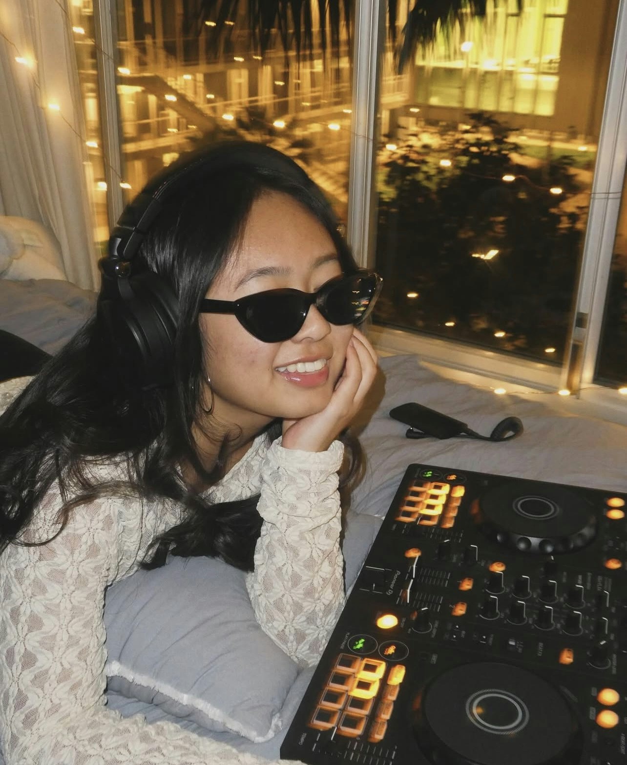 A young woman wearing sunglasses and headphones smiles while sitting next to a DJ controller, with a city backdrop at night.