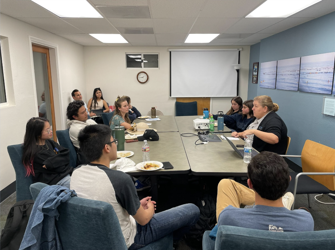 A group of people in a meeting room with a projector, eating and listening to a speaker.