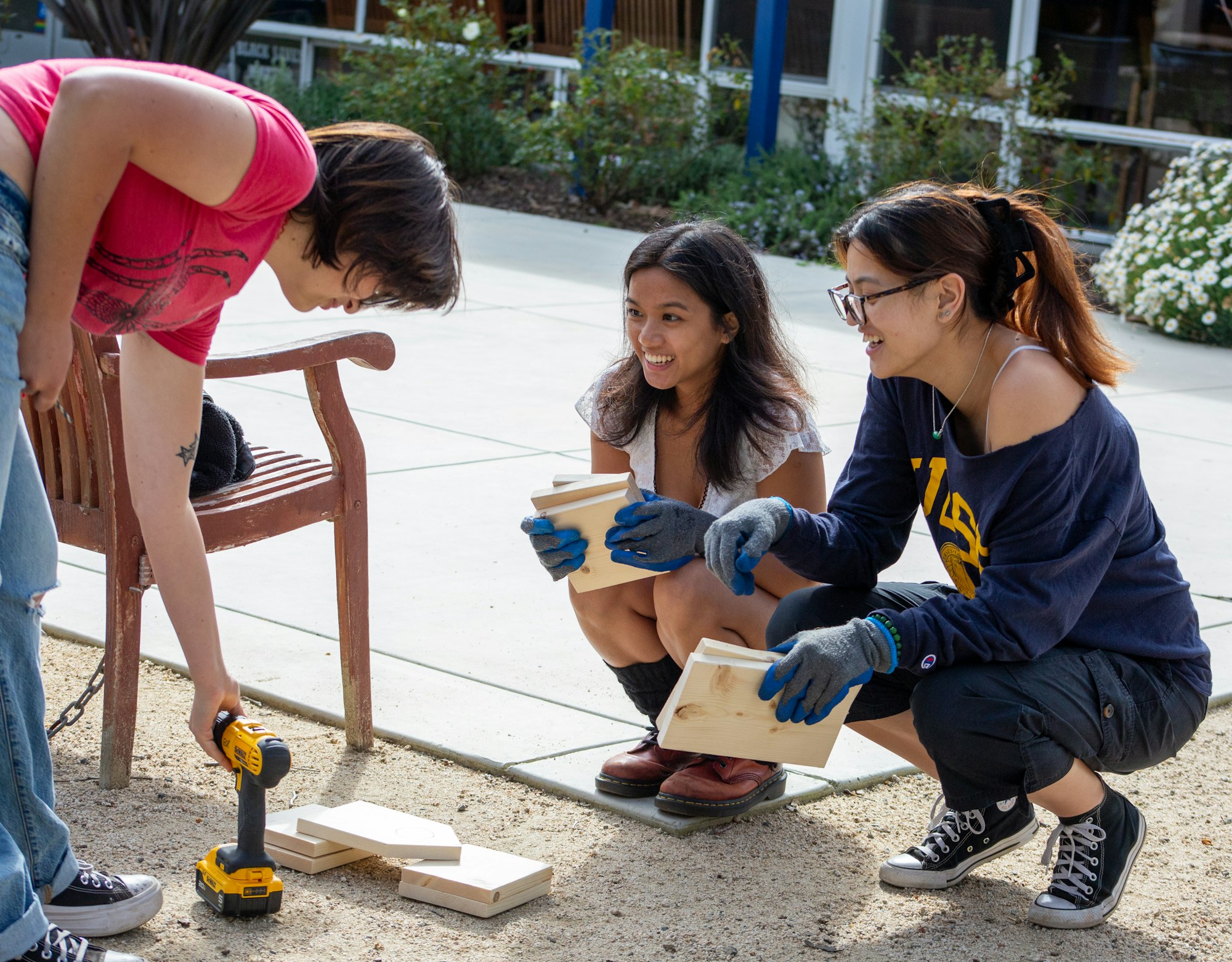 Three people outdoors with wood pieces, using a drill, and wearing gloves.