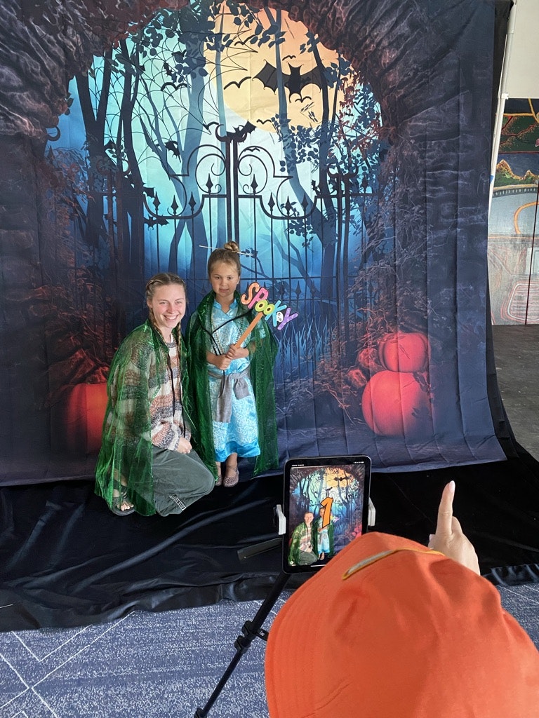 Two kids posing for a photo with a spooky forest backdrop. Someone is taking their picture using a smartphone on a tripod.