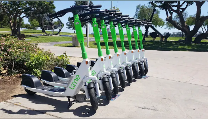 The image shows a row of electric scooters parked in a sunny outdoor area, likely available for rent.