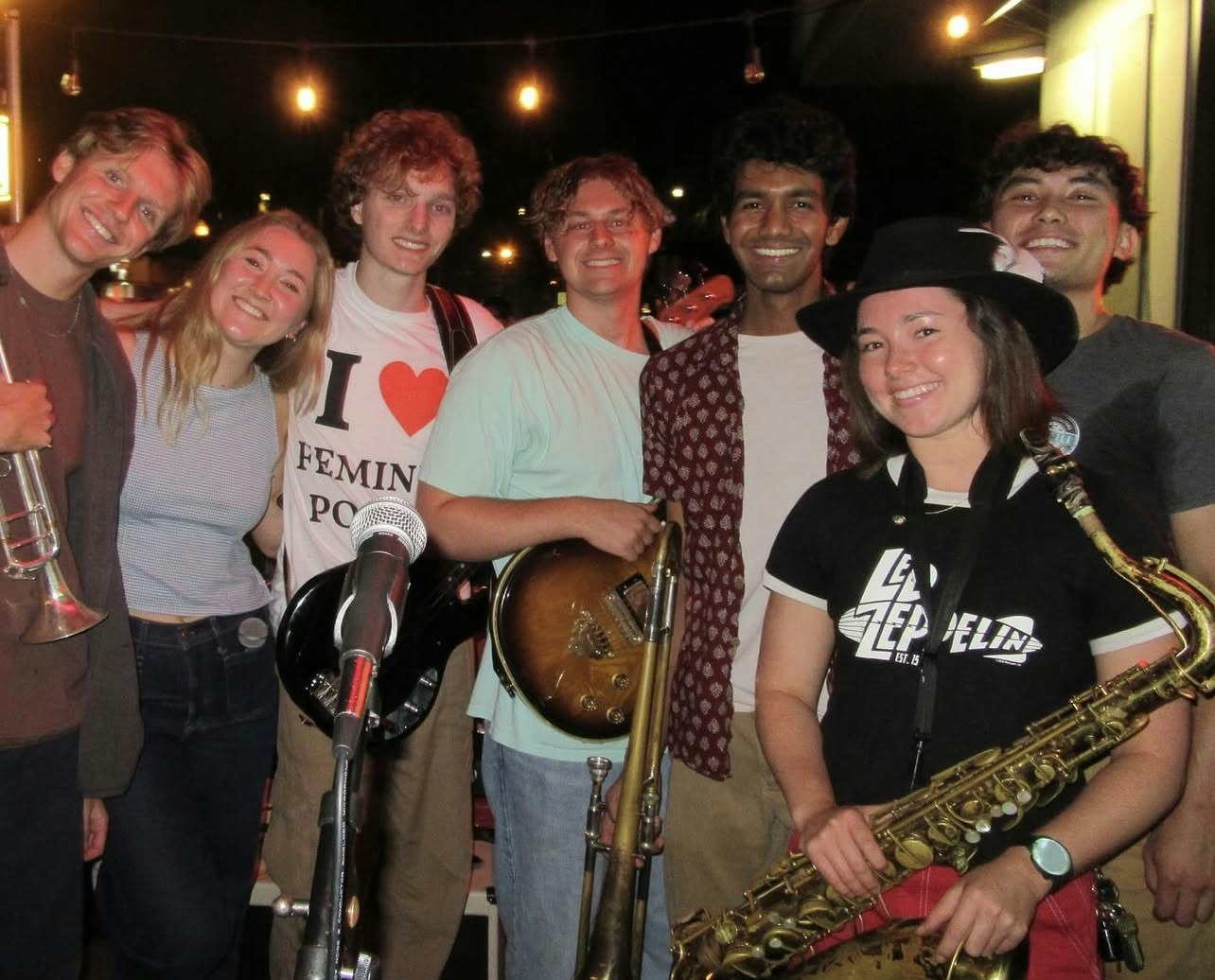 A group of eight young musicians poses together, smiling, with instruments, in a lively night setting.