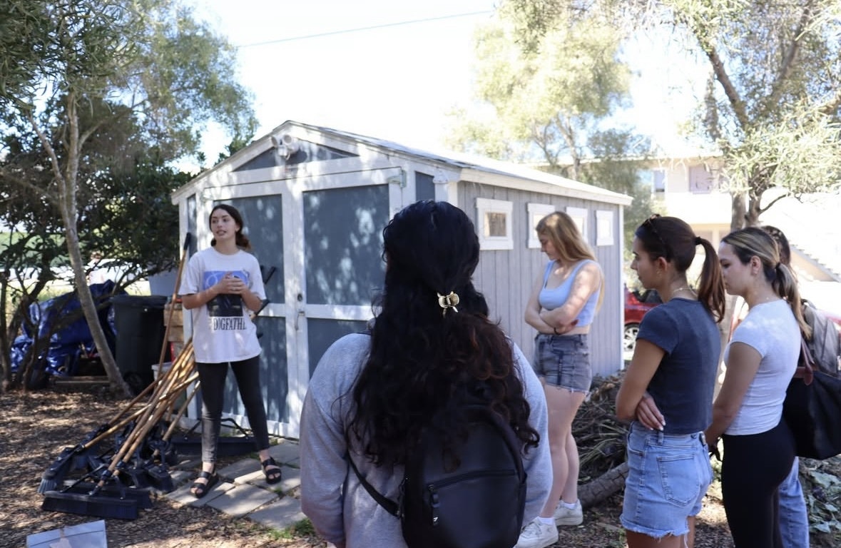 A person is speaking to a group near a shed, with tools visible outside.