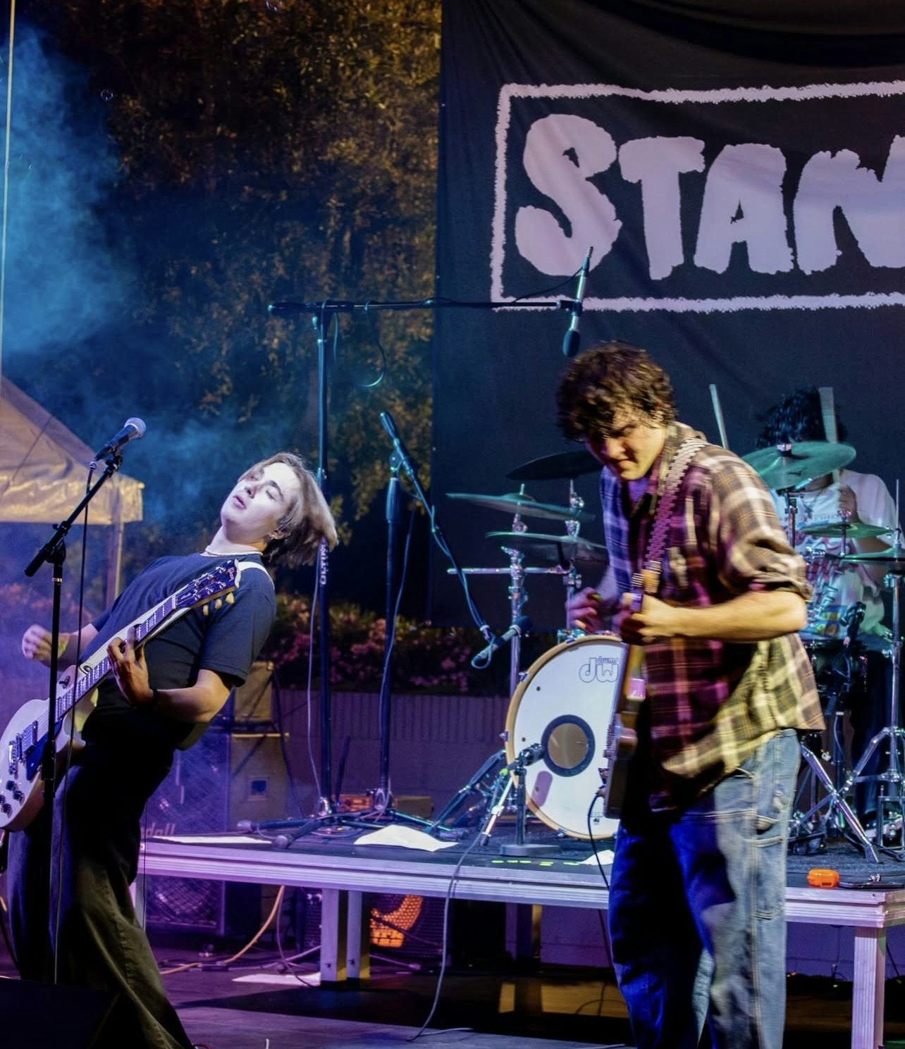 Two musicians perform on stage, with instruments and equipment, amidst smoke and a backdrop displaying the word "STAND."