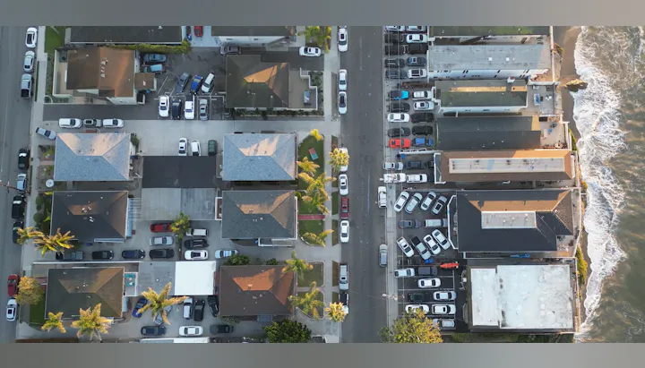 Aerial view of a coastal street with houses, parked cars, and waves hitting the shore.
