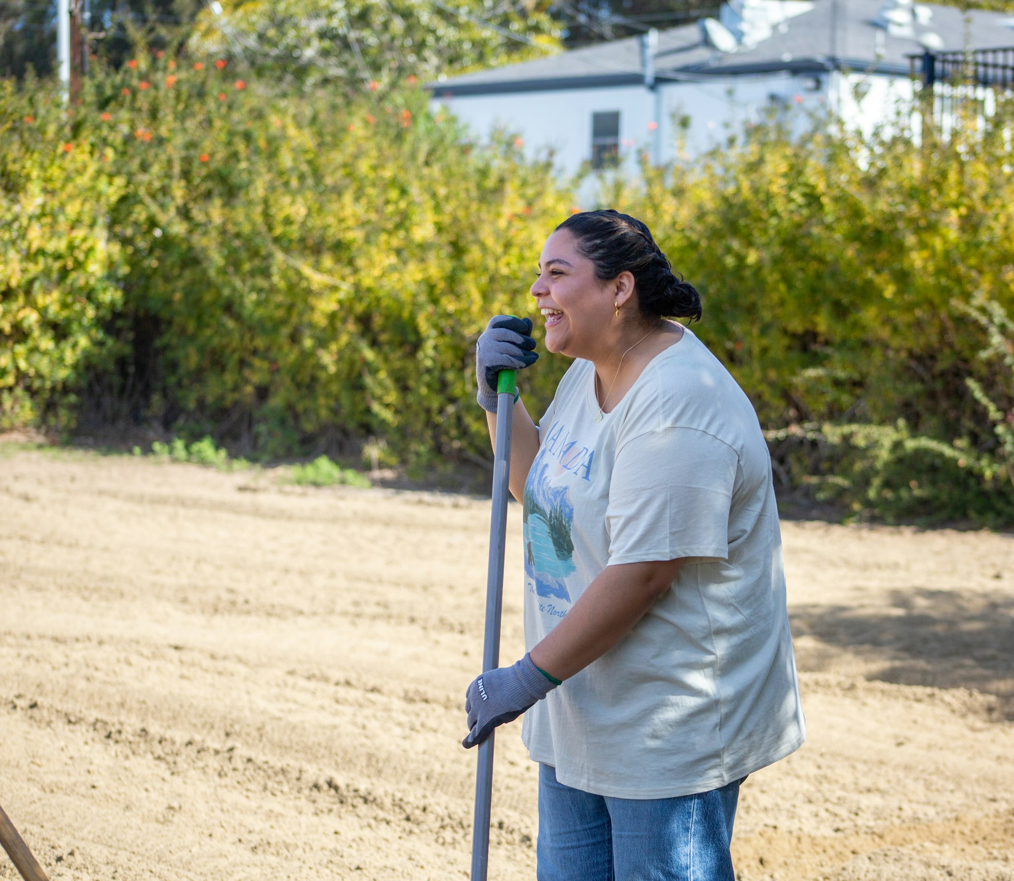 A person is smiling outdoors, holding a rake, wearing gloves, and standing on a dirt path with greenery and a white building in the background.