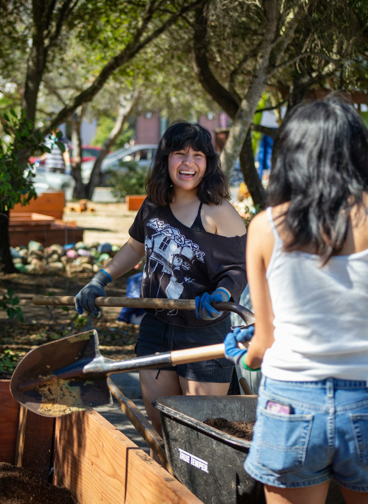 Two people gardening with shovels, smiling, wearing gloves, surrounded by trees.