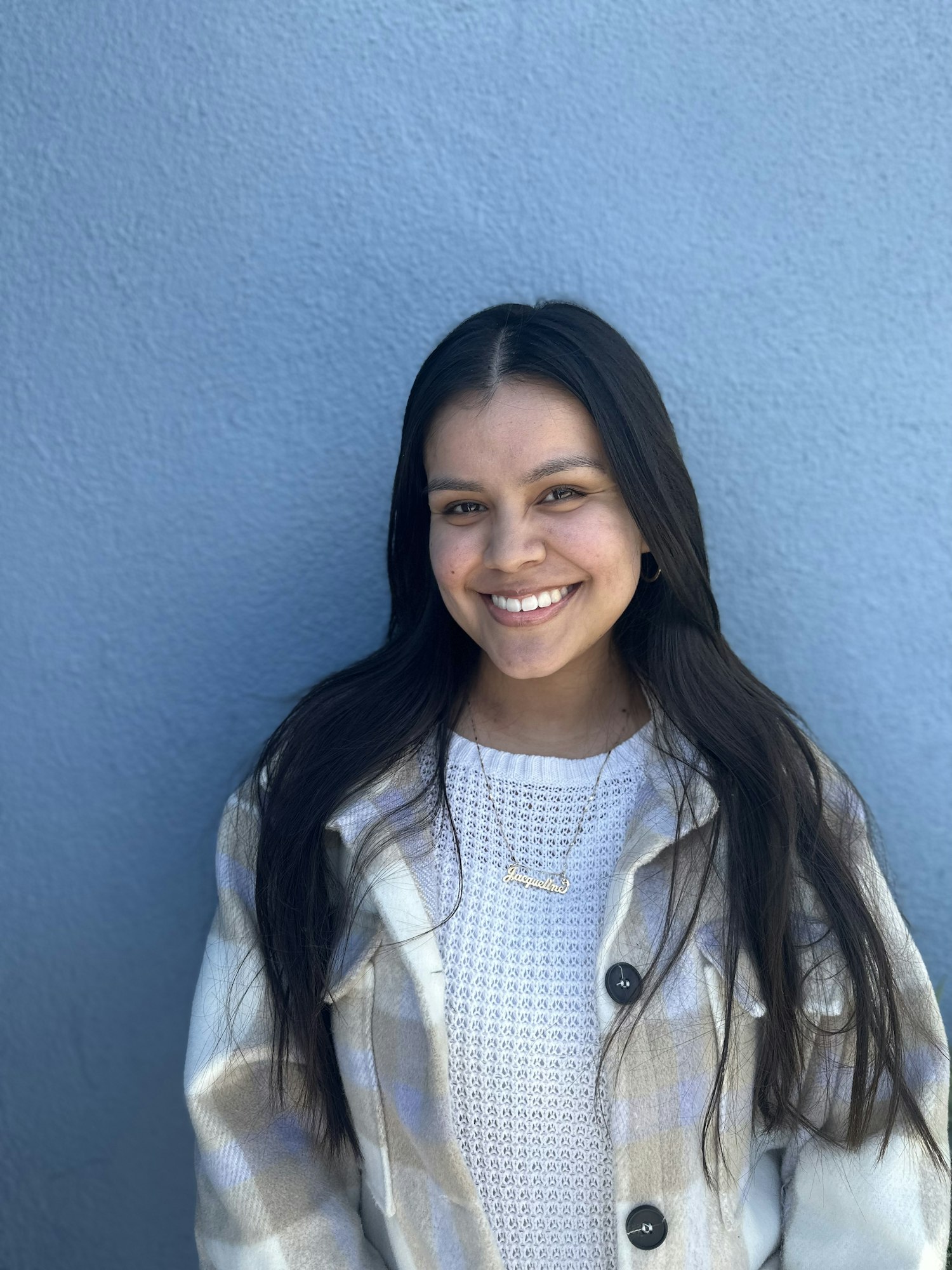 A smiling woman in a white patterned coat against a blue wall.