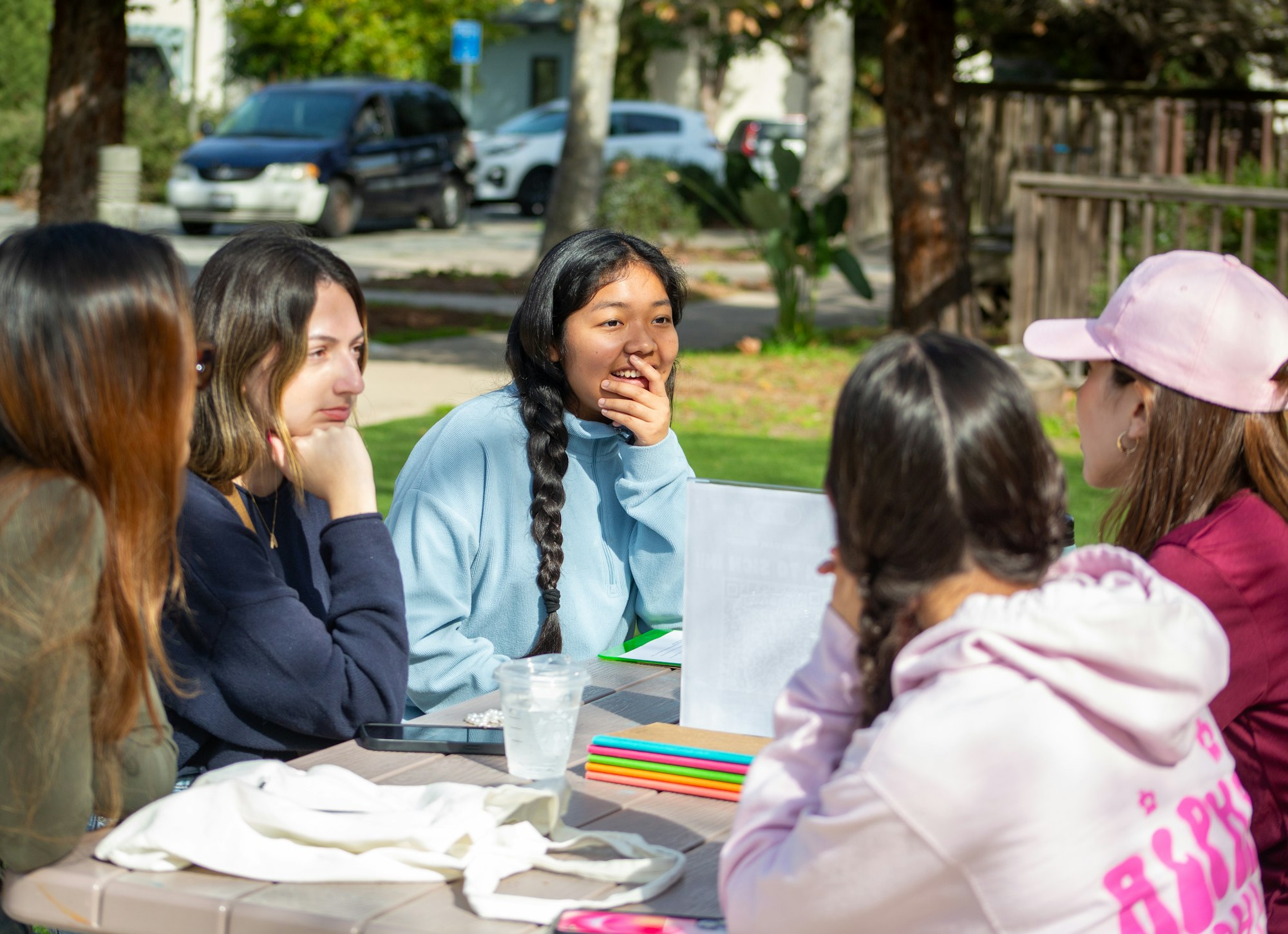 Five people sitting at an outdoor table, engaged in conversation, with notebooks and pens visible.