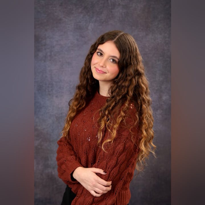 A person with long, curly hair wearing a brown sweater against a textured gray background.