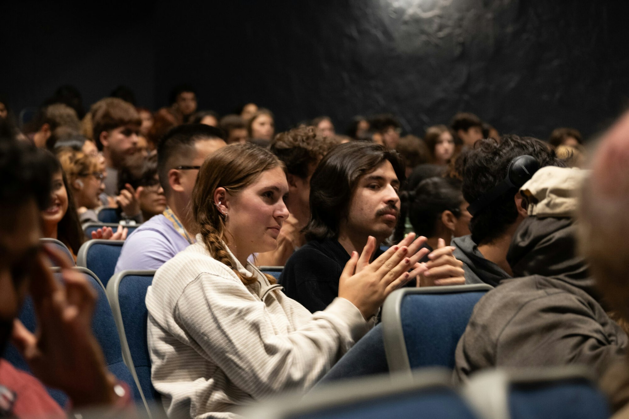 A crowded audience claps, with two individuals in focus. The setting appears to be a seminar or presentation.