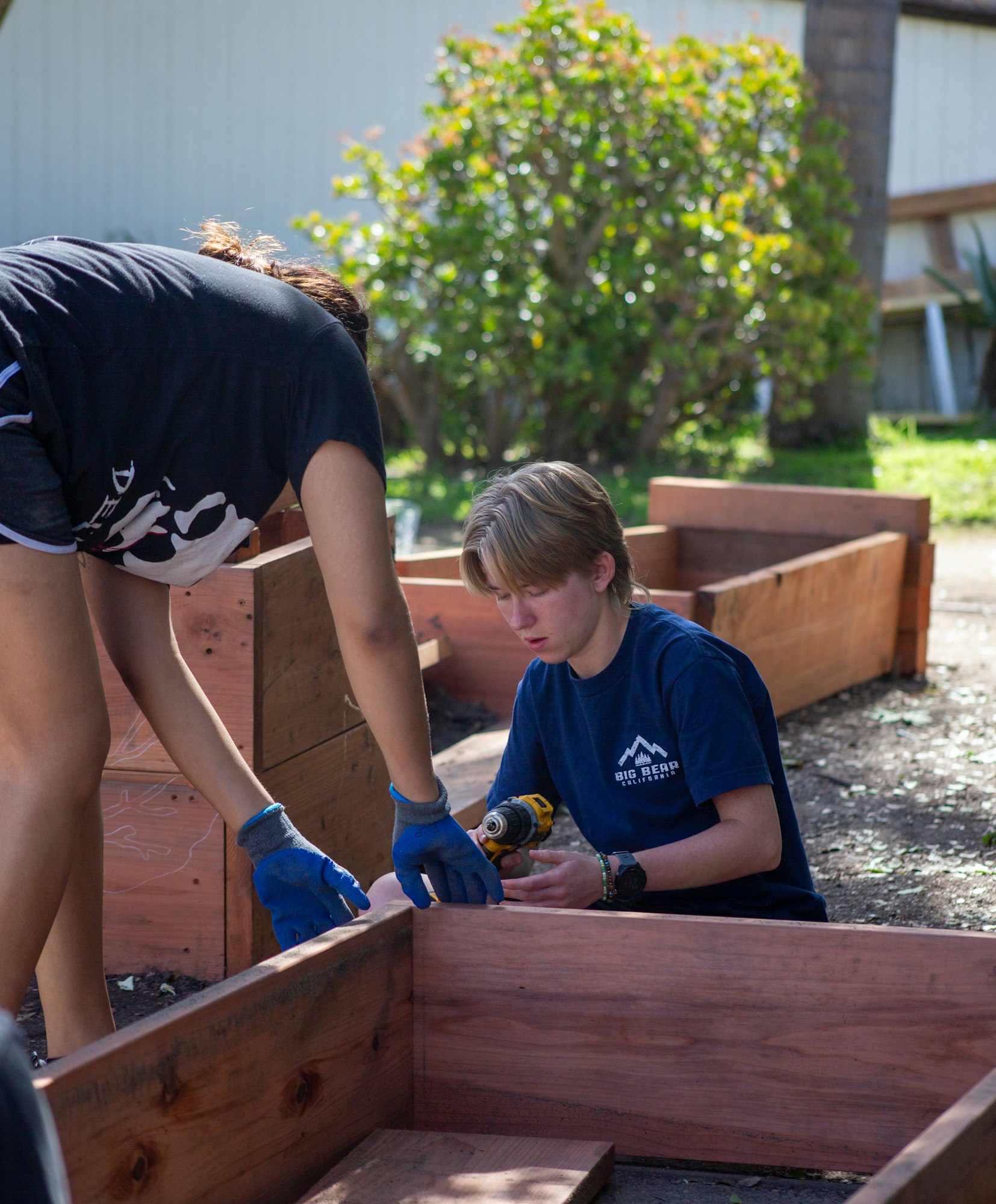 Two people are working together outdoors, assembling wooden structures with a drill.