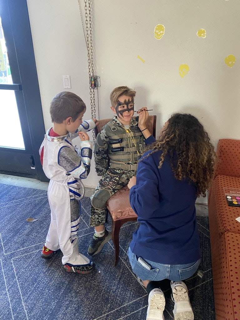 Two children in costumes with one getting face paint by an adult.