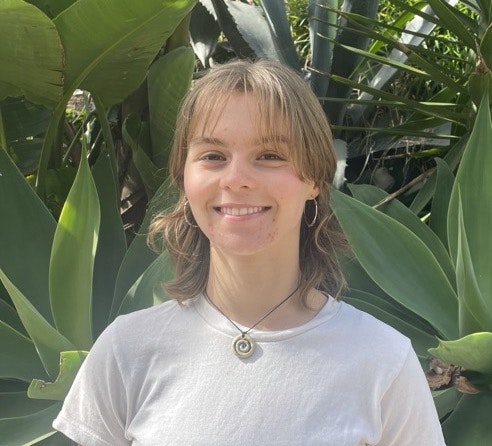 A smiling person with a white shirt and a pendant necklace, standing in front of green foliage.