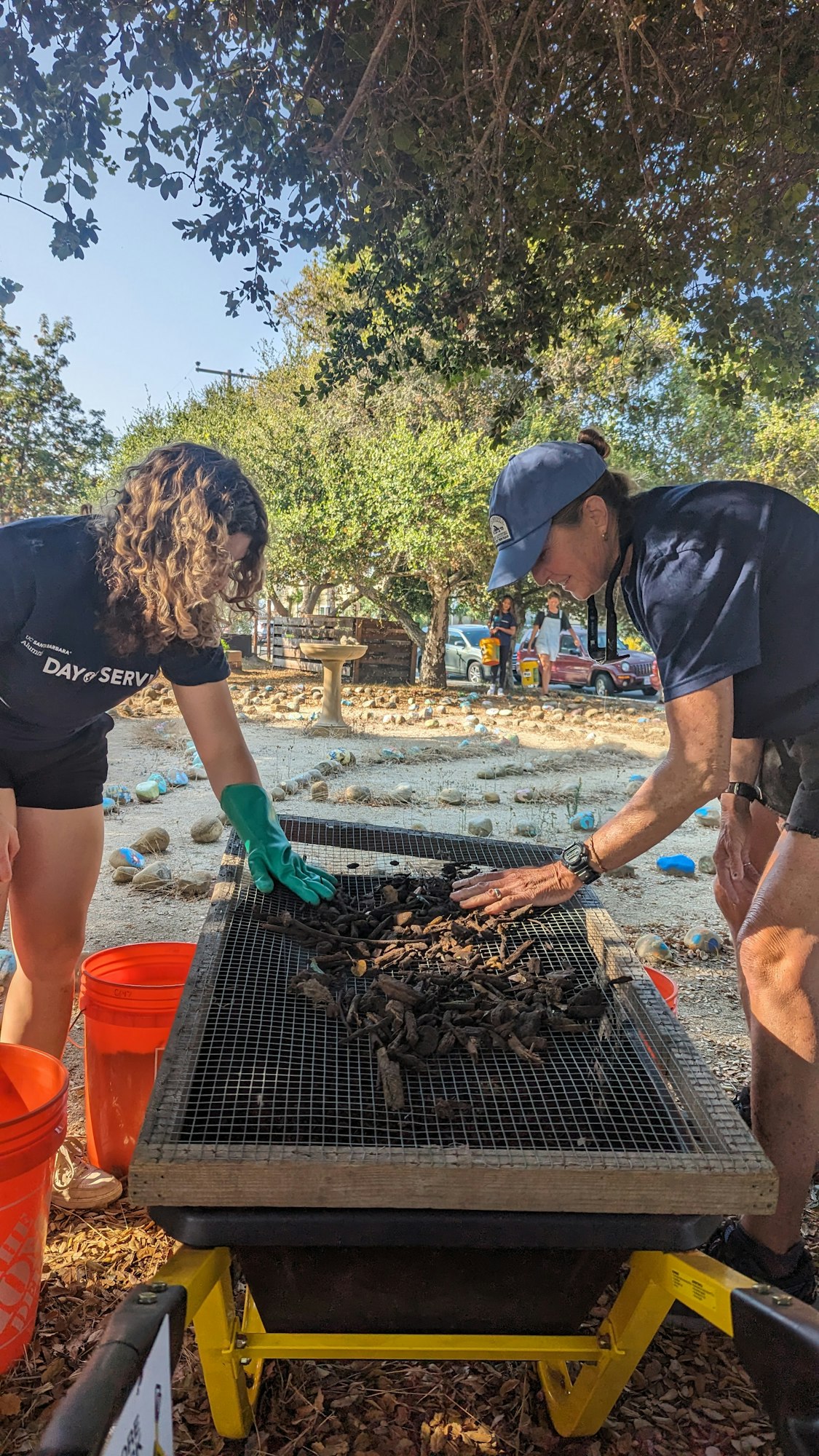Two people are sorting debris on a mesh screen outdoors, with buckets nearby.