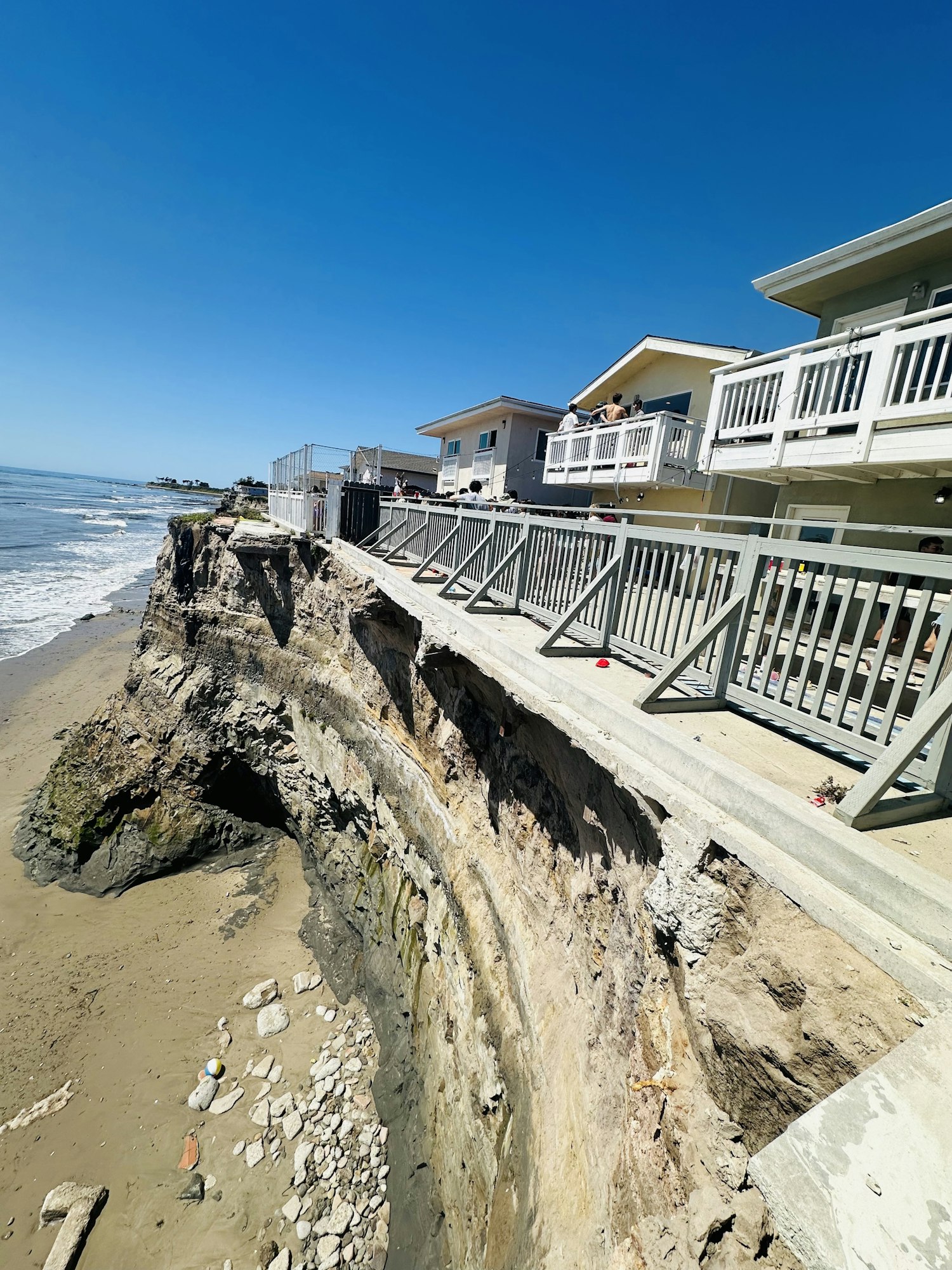 A cliffside houses with a boardwalk overlooking a beach, showing erosion and exposed rocks near the shore. Clear blue sky above.
