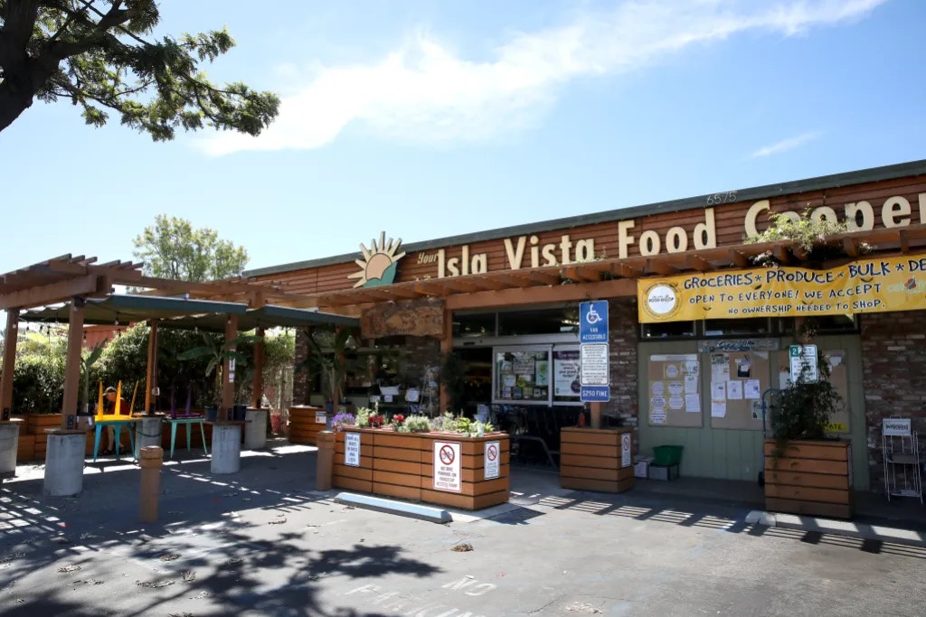 The image shows the exterior of Isla Vista Food Cooperative, featuring a welcoming entrance, outdoor seating, and greenery.