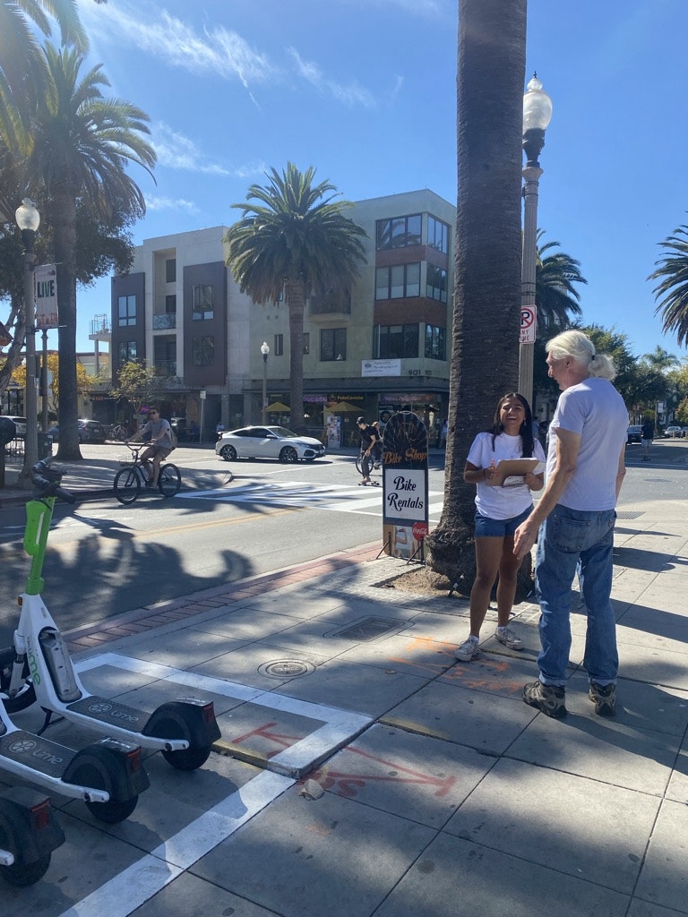 May contain: street, city, urban, road, palm tree, tree, plant, path, pants, clothing, neighborhood, teen, girl, female, person, e-scooter, vehicle, transportation, sidewalk, jeans, car, shorts, bicycle, shoe, footwear, wheel, and machine