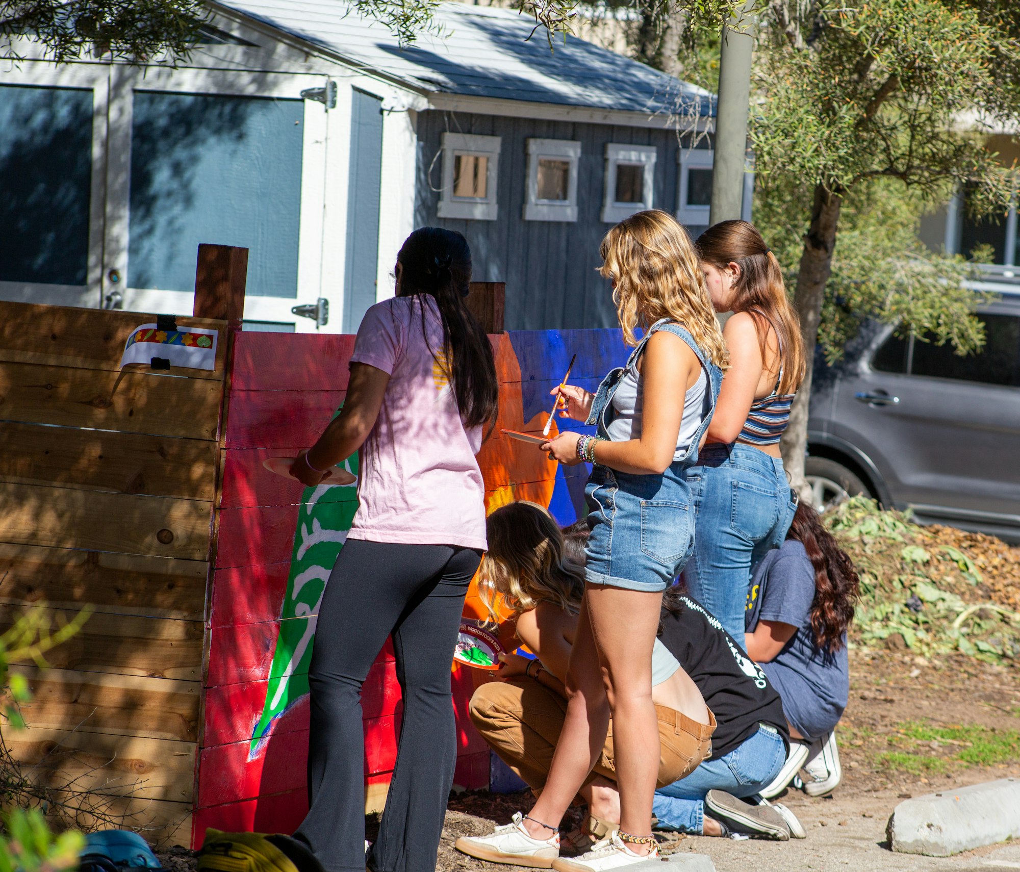 A group of people painting a wooden fence with colorful designs outdoors near a building.