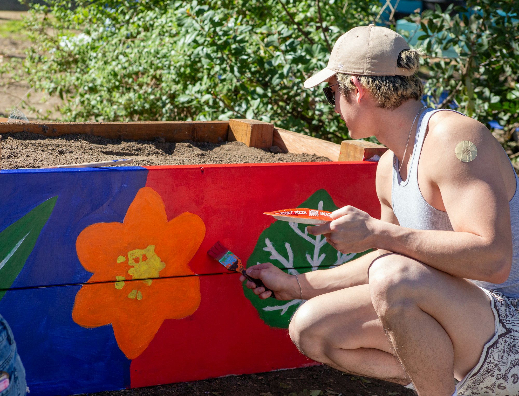 A person painting a colorful flower mural on a wooden garden box outdoors.