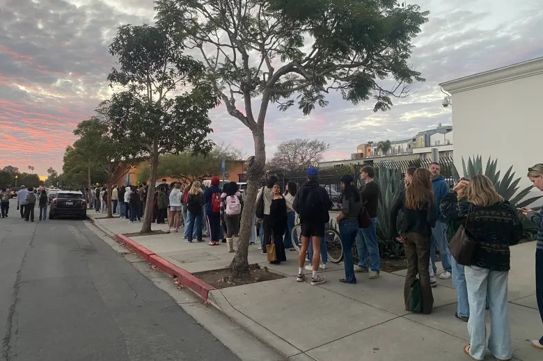 A long line of people waiting outside, with trees and a colorful sky at sunset in the background.