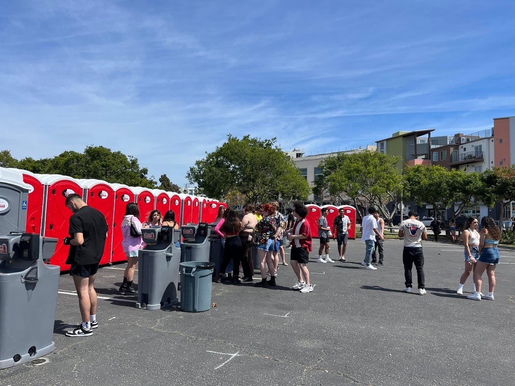 A sunny day with people lined up to use a row of portable toilets in a parking lot.