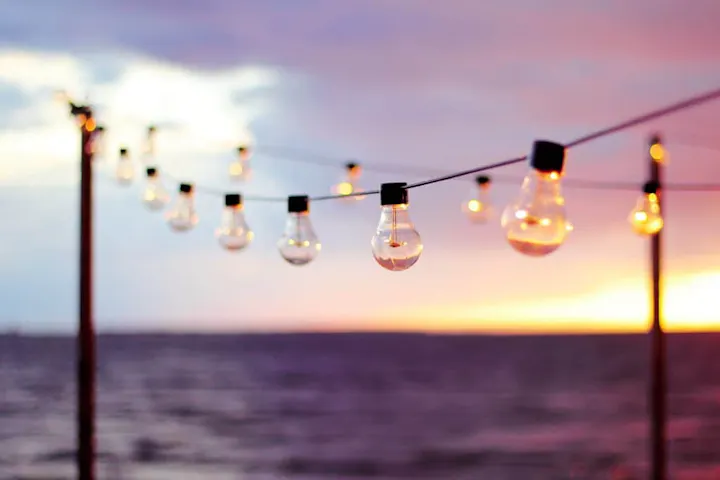 The image features a series of glowing light bulbs strung along a wire, against a colorful sunset over the water.