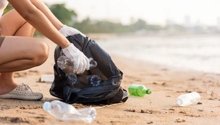 Person picking up litter on a beach into a black trash bag, with scattered plastic bottles around.