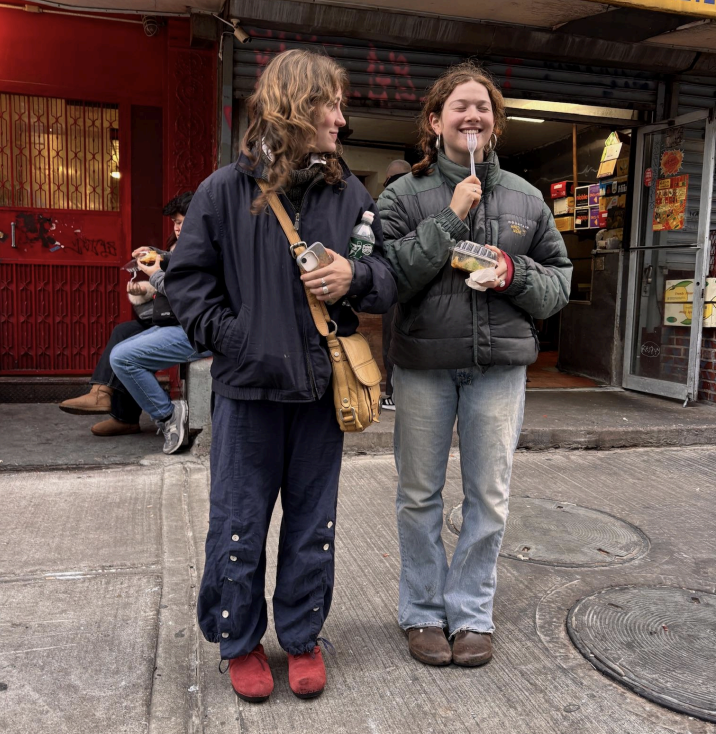 Two young people stand on a city street, smiling, one holding a snack and the other a phone. A shop and people are in the background.