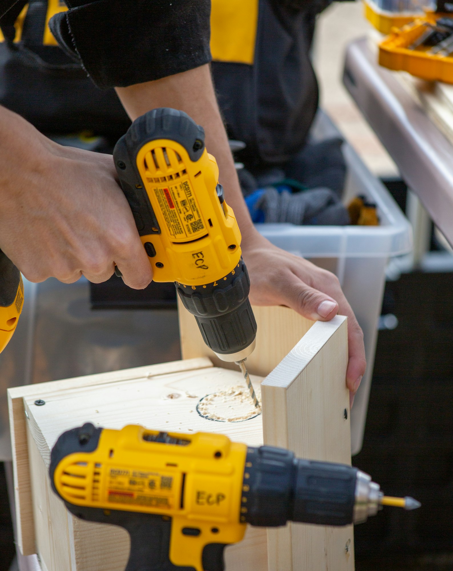 Person drilling a hole in wood with a yellow power drill, another drill is visible on the side.