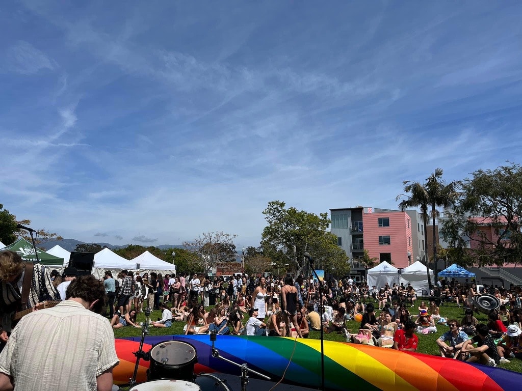 Outdoor event with a crowd gathered around a stage with drums, under a clear blue sky. A rainbow flag is visible in the foreground.