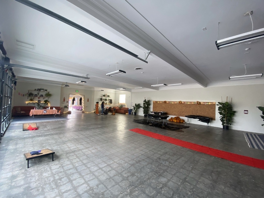 Indoor space with tables, chairs, plants, and people in the background; resembles a community hall or event space.