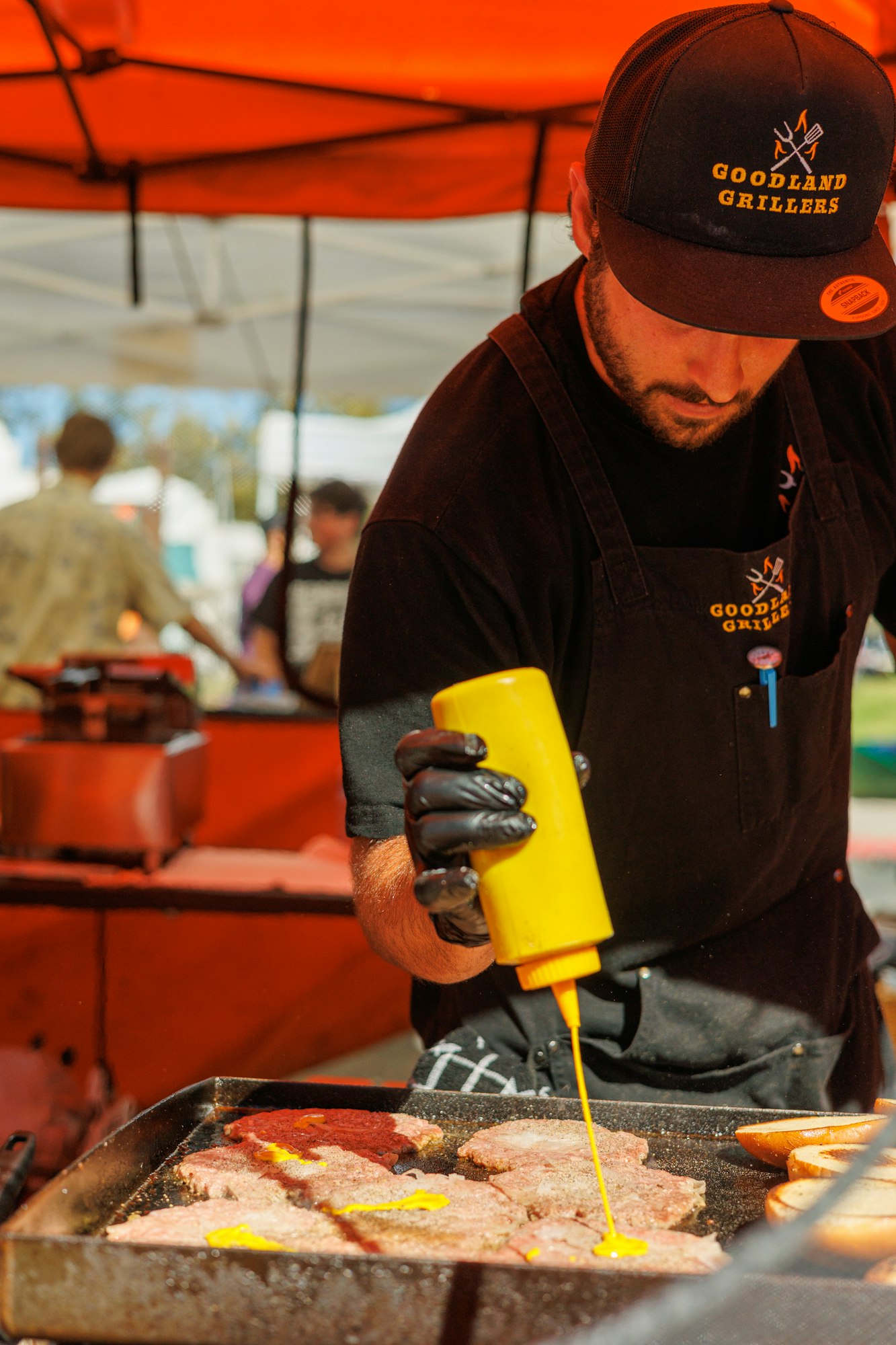 A person grilling meat at an outdoor event, applying sauce from a yellow squeeze bottle.