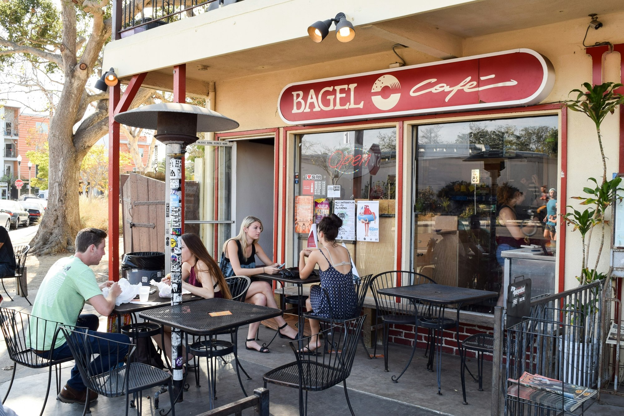 A cozy bagel cafe with outdoor seating, where people are enjoying their meals and chatting in a sunny environment.
