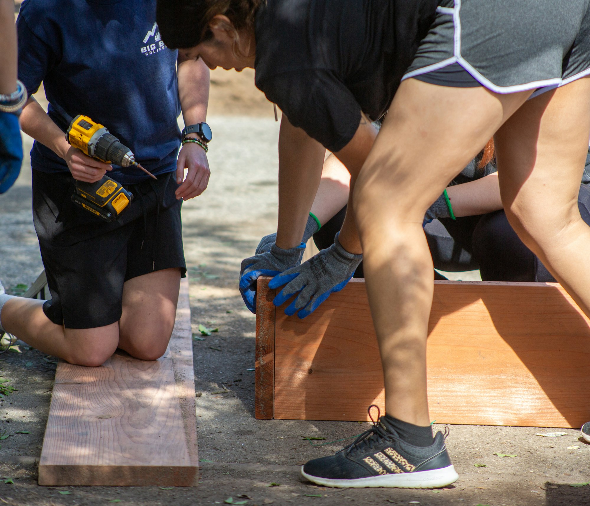 People working outdoors with wood, one holding a drill, others wearing gloves and assembling boards.