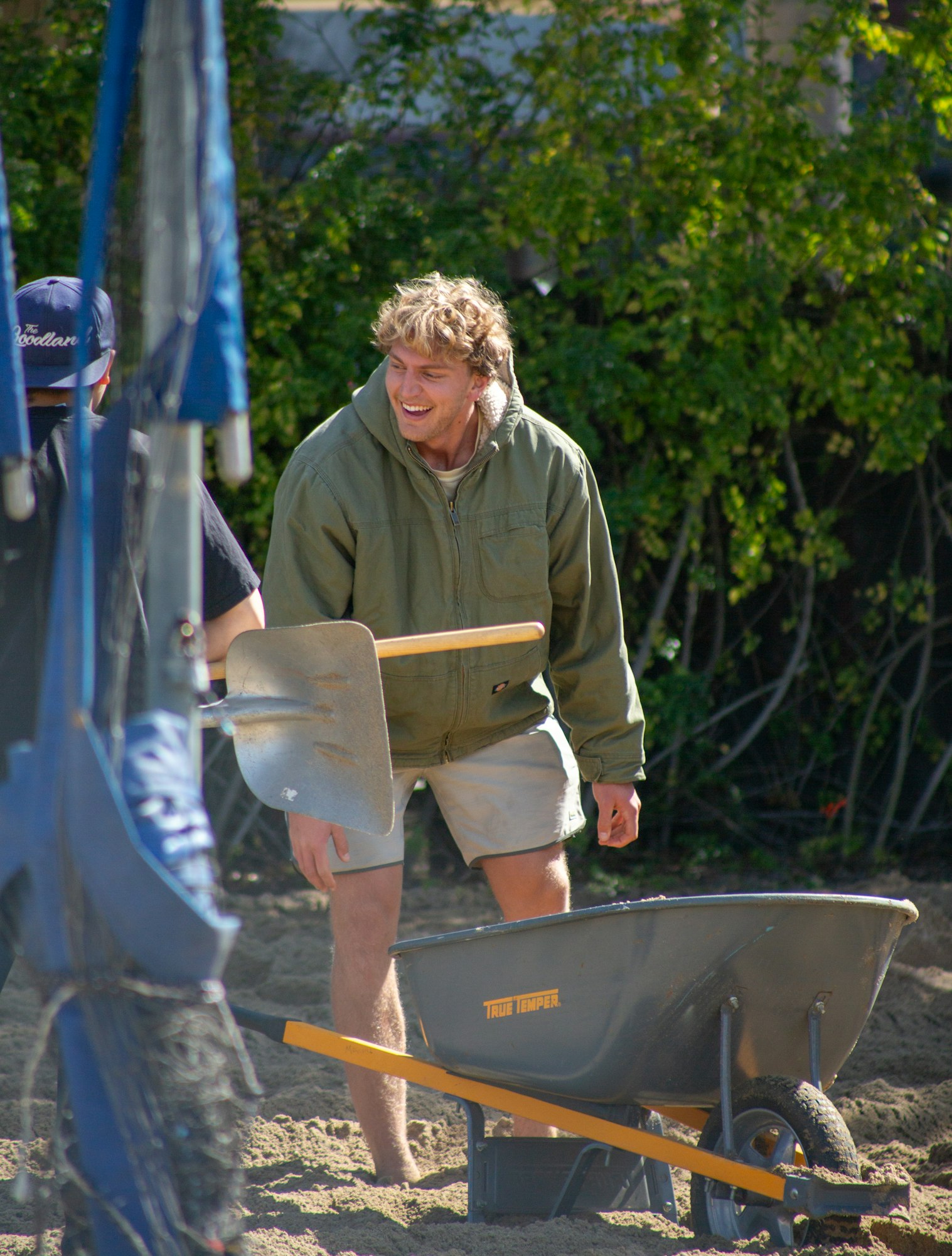 A person outdoors, wearing a green jacket, stands near a wheelbarrow and a shovel. They're smiling, with greenery in the background.