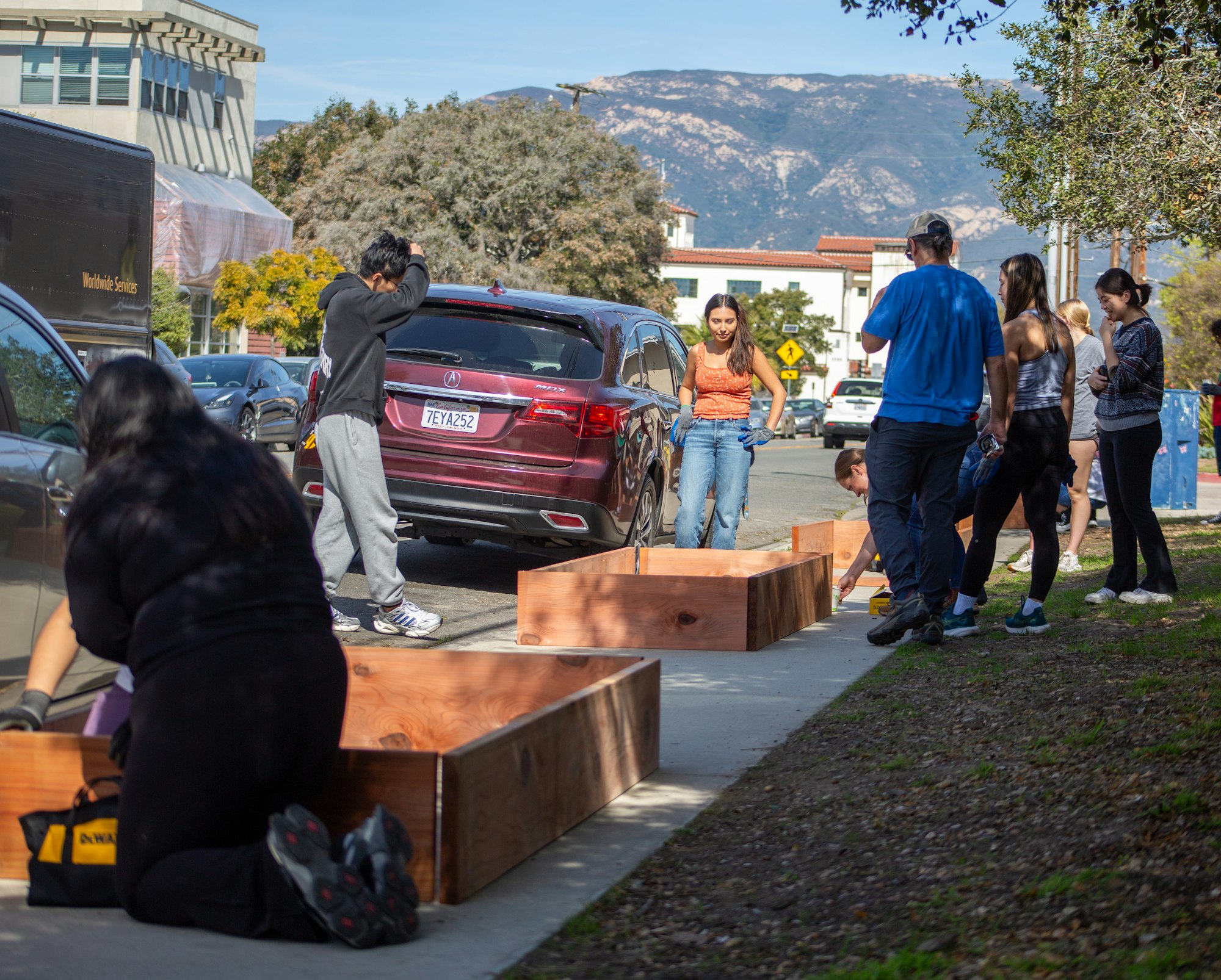 People assembling wooden boxes on a sidewalk near parked cars.