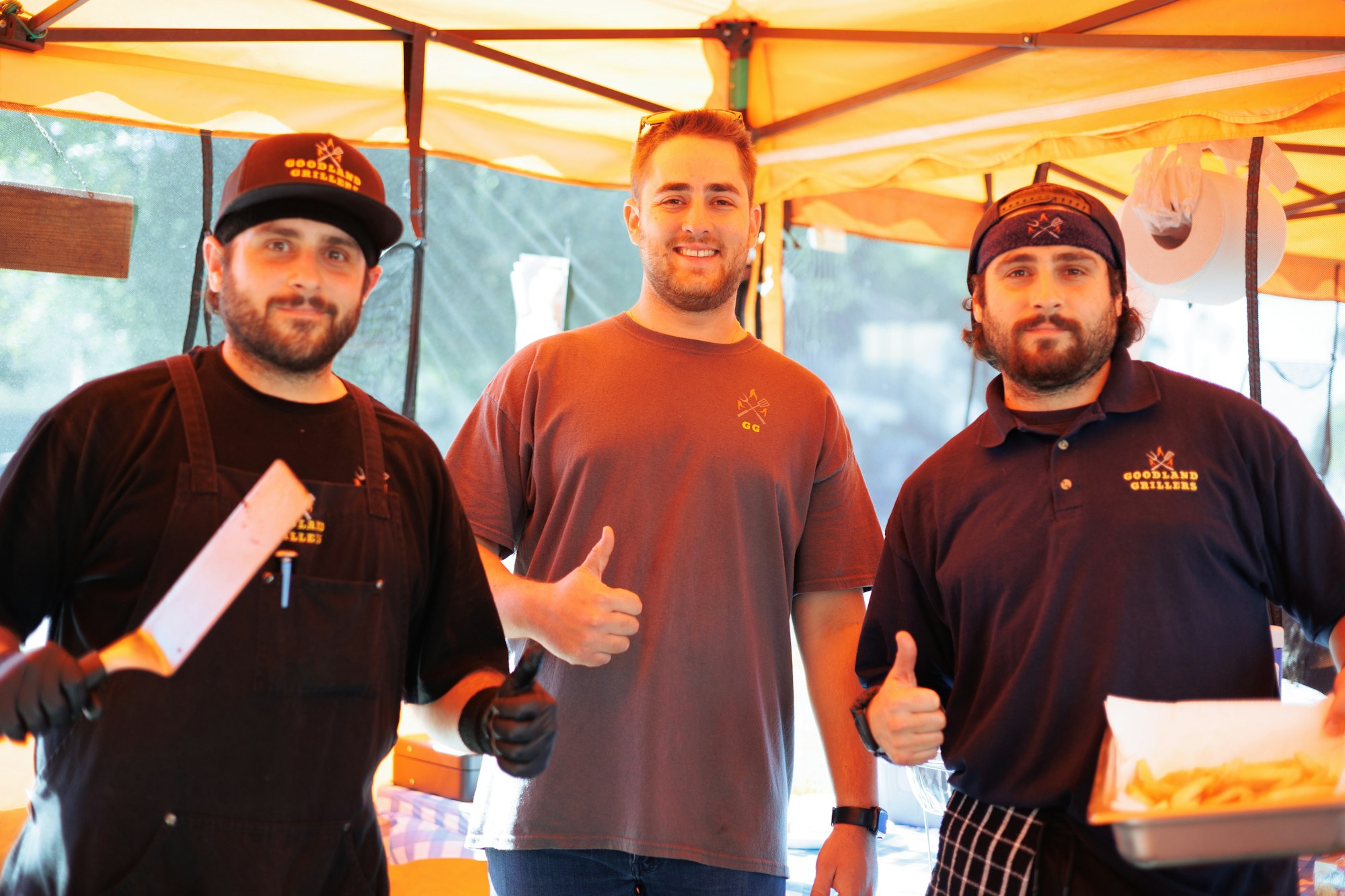 Three men smiling at a food stall, two in chef attire and one holding a cleaver; "Goodland Grillers" logo visible.