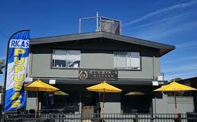 The image shows a restaurant with a sign for "Rica's Pupusas," featuring outdoor seating with yellow umbrellas.