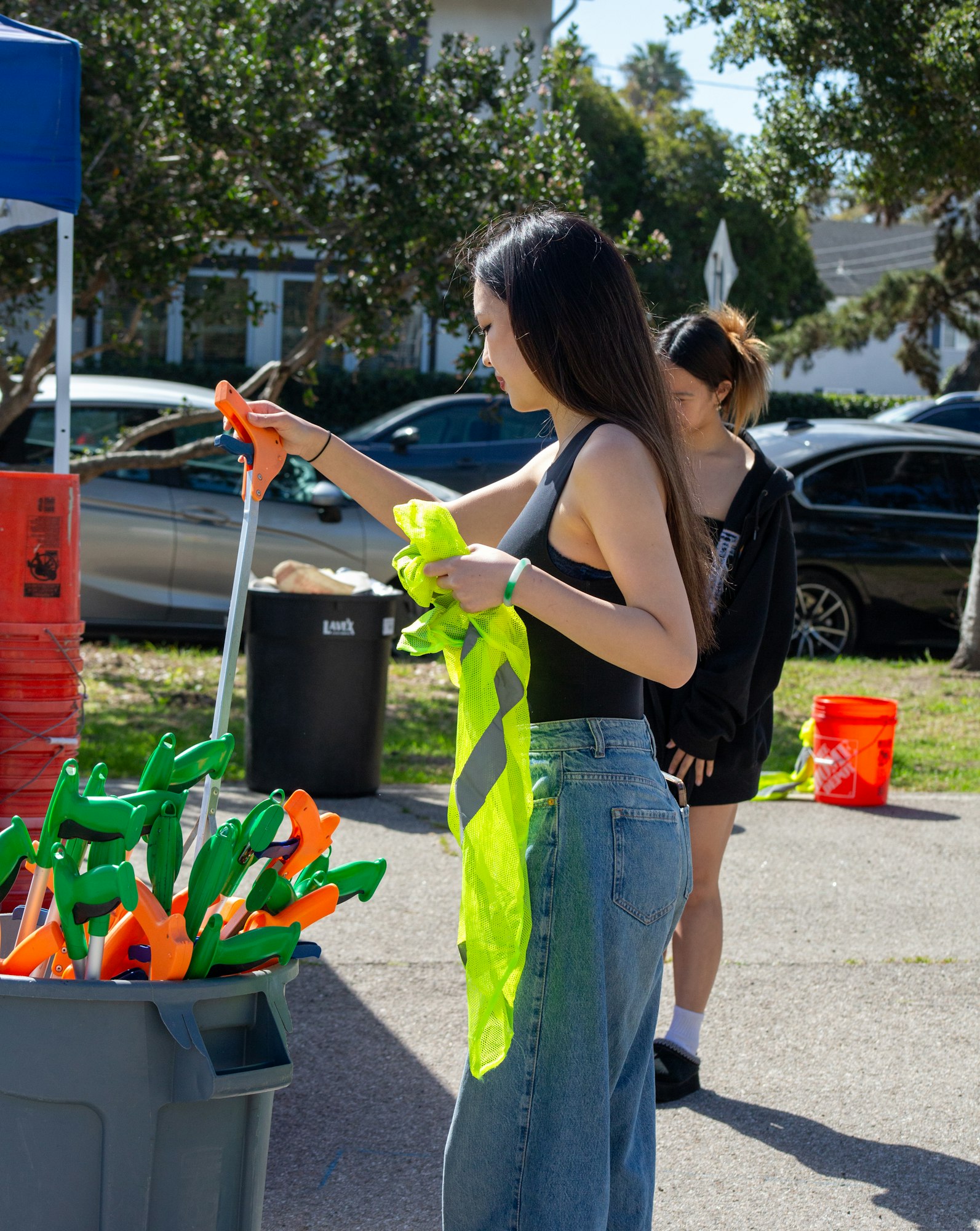 Two people are outside preparing for an activity with grabbers and safety vests near a bin.