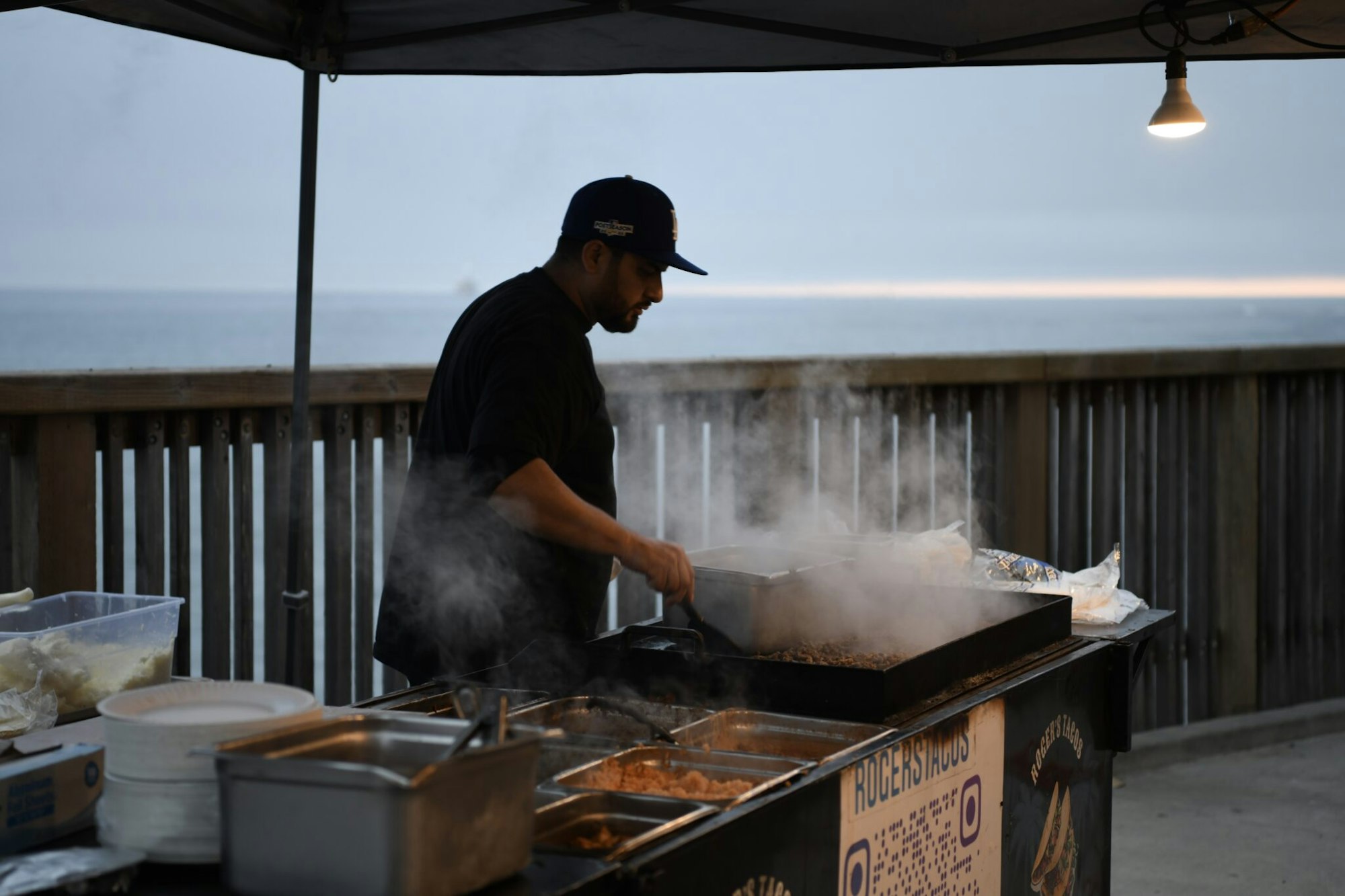 A chef is cooking at a food stall by the water, with steam rising from the grill and a coastal view in the background.