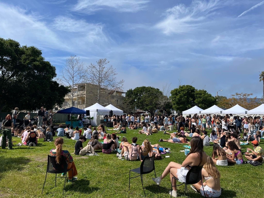 Outdoor gathering with seated people, tents, and a band performing under blue skies.