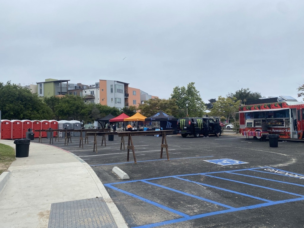 Outdoor event setup with food trucks, portable toilets, tables, and canopies in a parking lot.