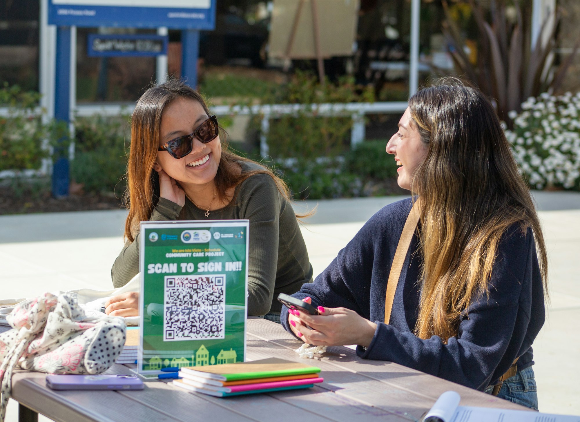 Two people are smiling and talking at a table outdoors. A sign with a QR code is on the table.